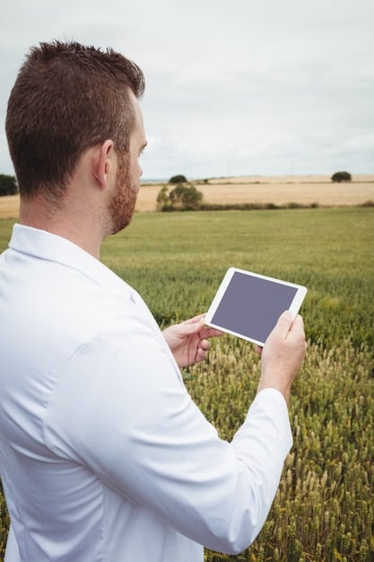 A farmer using a tablet in a field to monitor crop health. The tablet displays data visualizations showing soil moisture levels, nutrient content, and plant health metrics. The farmer is wearing a hat and work clothes, indicating a hands-on approach to data-driven farming.